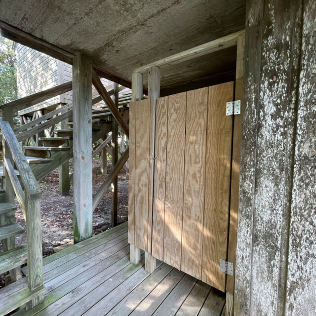 Outdoor wooden shower stall under stairs next to a concrete wall with visible wood and metal hinges.