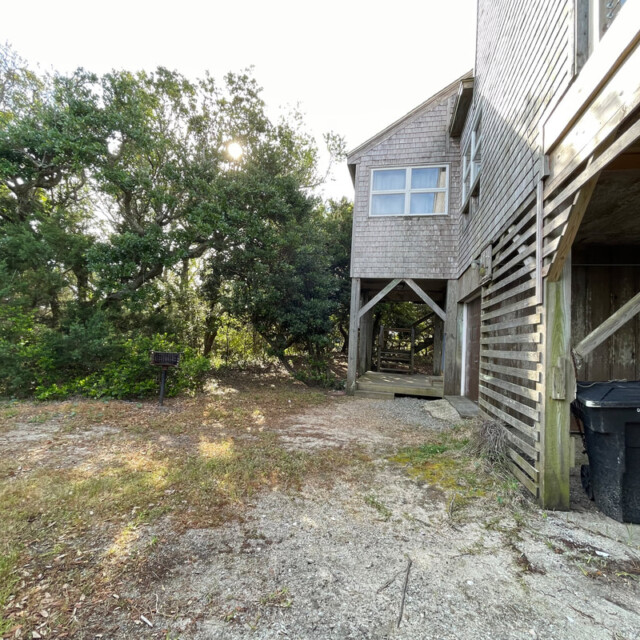 A two-story wooden house with a gravel yard, trees, and a trash bin under an open deck.