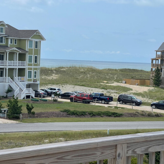 Large beach houses and parked cars near grassy dunes with the ocean visible in the background under a clear sky.