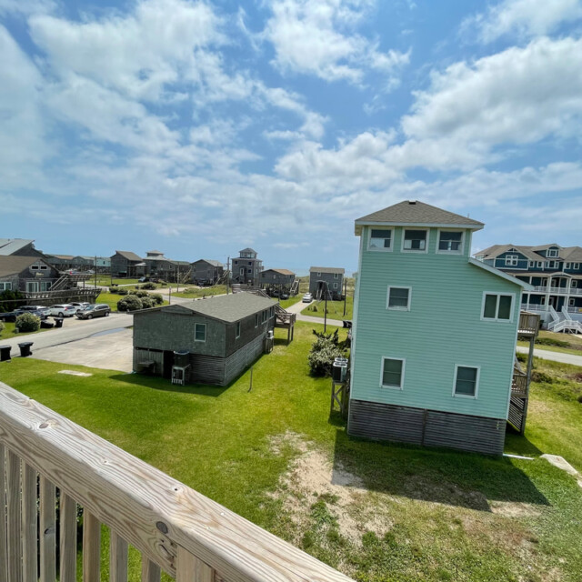 View of coastal houses with green lawns and a partly cloudy sky, taken from a wooden balcony overlooking the neighborhood.