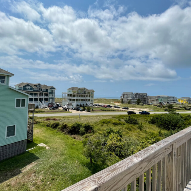 View from a wooden deck overlooking houses, parked cars, grass, and a partly cloudy sky near a beach.