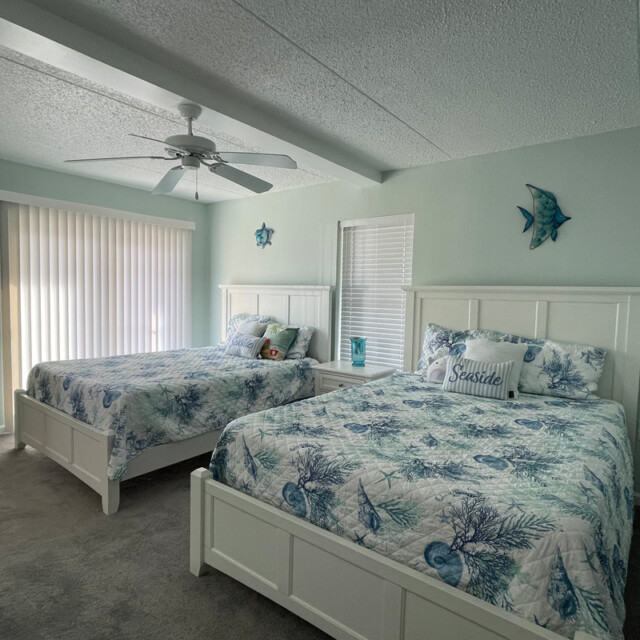Bedroom with two white beds, ocean-themed bedding, a ceiling fan, wall art, and sunlight from a window and sliding door.