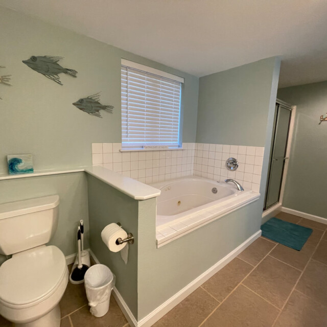Bathroom with toilet, whirlpool tub, wall art of fish, tiled floor, shower, window with blinds, and wall hooks.