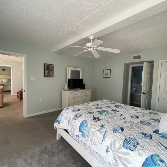 Bedroom with a seashell-patterned bedspread, dresser with TV, ceiling fan, and open doorways to other rooms.