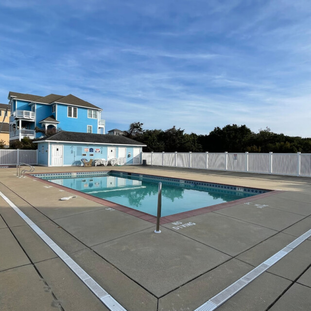 Outdoor swimming pool with fence, pool chairs, and a blue house in the background under a clear sky.