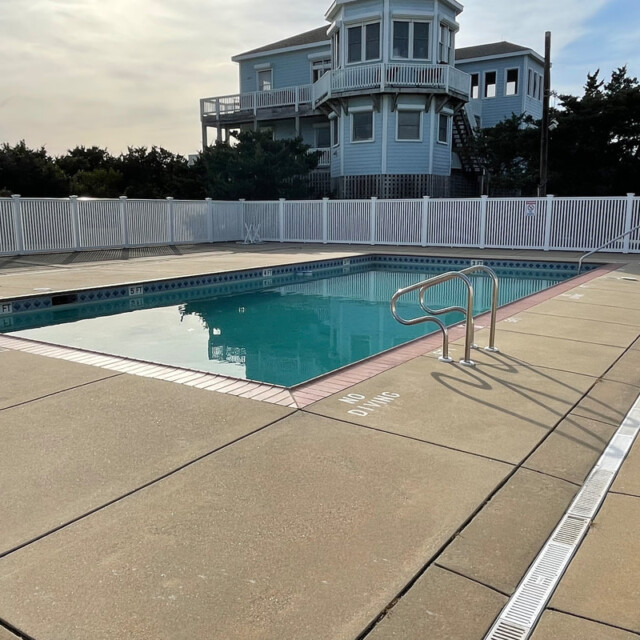 Empty outdoor swimming pool with handrails, surrounded by a fence, and a large house in the background.