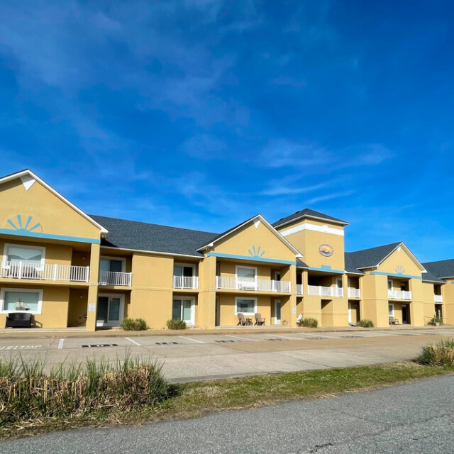 Two-story yellow motel building with balconies, white trim, and an empty parking lot under a clear blue sky.