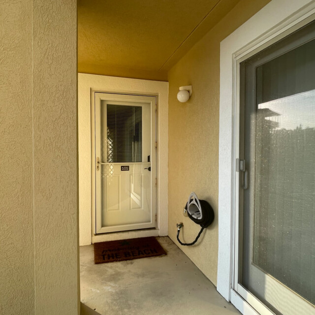 Small porch with a white door, wall light, coiled hose, and a doormat that says “THE BEACH.”.