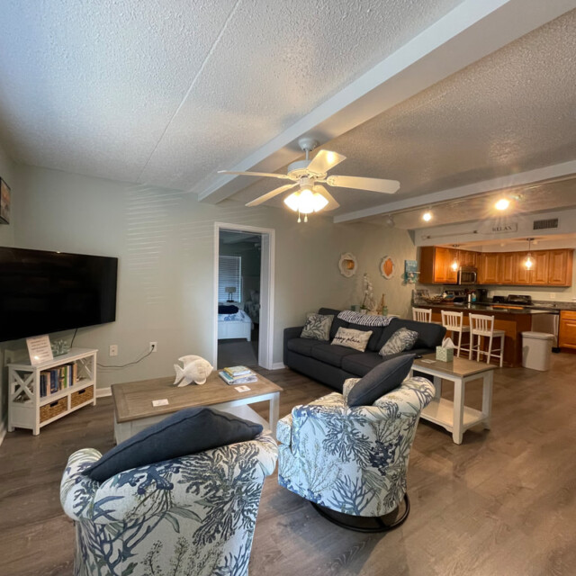 Living room with two armchairs, a sofa, a TV, and view into a kitchen and dining area with wooden cabinets and table.