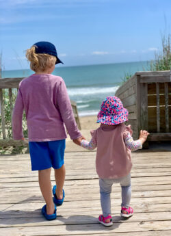 Two young children walk hand in hand on a wooden boardwalk toward the beach on a sunny day.