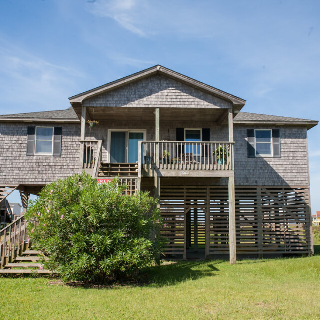 Gray wooden beach house on stilts with a front porch, stairs, and a green bush in the yard under a clear sky.