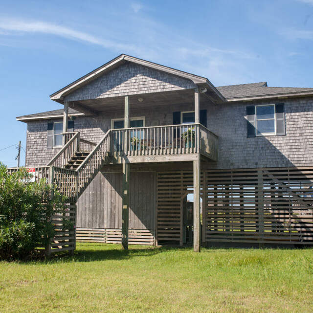 A gray wooden beach house on stilts with a front porch, stairs, and grassy yard under a clear blue sky.