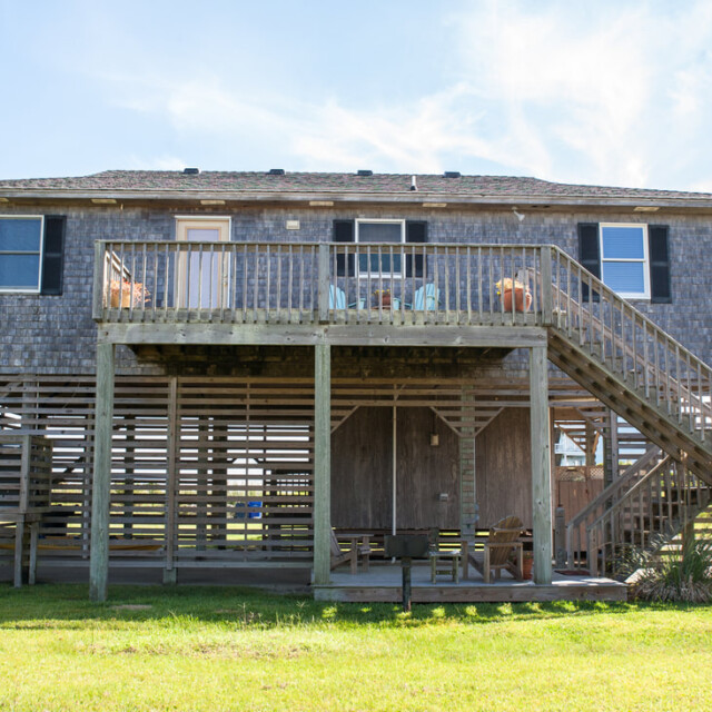 Elevated wooden house with a large front porch, exterior stairs, and an open area underneath, on a sunny day.