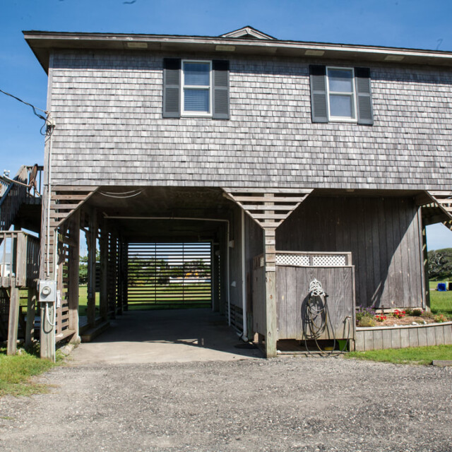 Elevated wooden house with exterior stairs, gravel driveway, and open ground level on a sunny day.