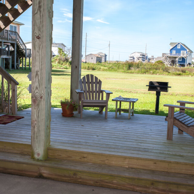 Wooden patio with two chairs, a small table, potted plants, and a grill, overlooking a grassy area and houses.