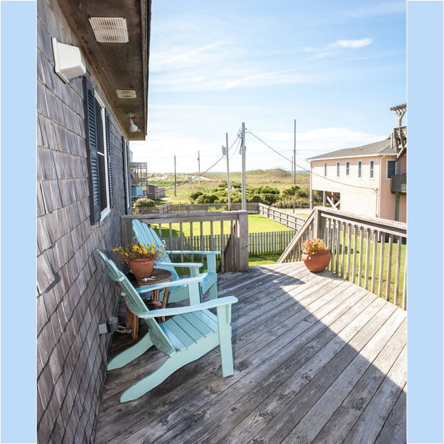 Two light-blue Adirondack chairs and potted flowers on a wooden deck overlooking grass and distant dunes.
