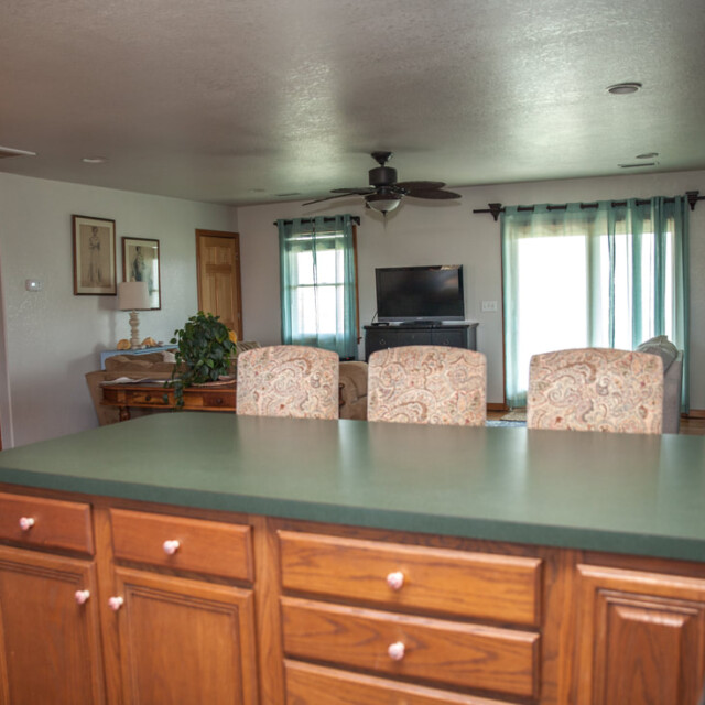 Open kitchen with green countertop, wood cabinets, and view into a living room with TV, ceiling fan, and windows.