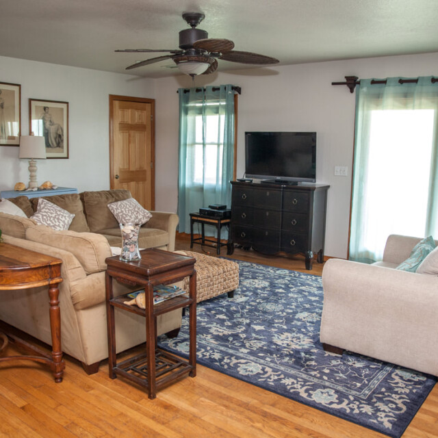Living room with two sofas, TV on a black dresser, blue patterned rug, wooden floors, and large windows with sheer curtains.
