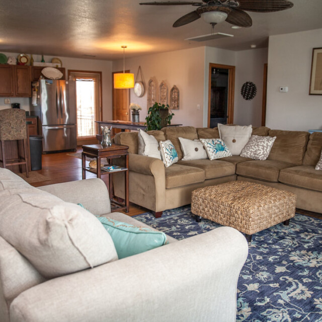 Open living room with two beige sofas, a wicker coffee table, blue rug, and a kitchen in the background.