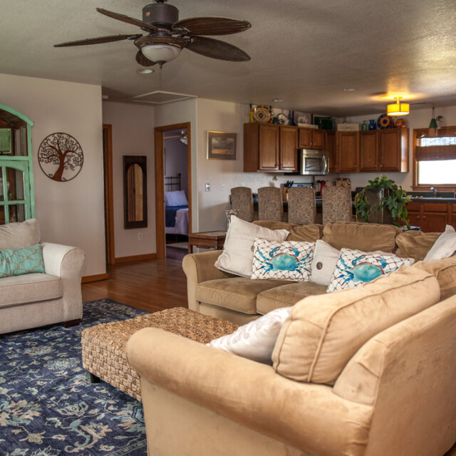 Living room with tan couches, blue rug, and open kitchen in the background with wood cabinets and stainless steel fridge.