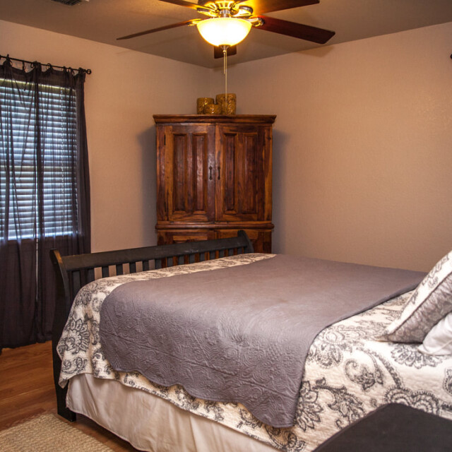 Bedroom with a wooden bed, patterned bedding, wooden armoire, two windows with dark curtains, and a ceiling fan.
