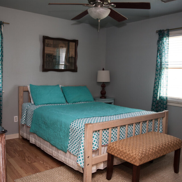 Bedroom with teal bedding, wooden bed frame, nightstand, lamp, mirror, dresser, and two windows with teal curtains.