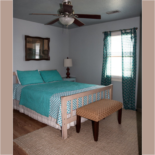 Bedroom with a turquoise bedspread, wooden bench, ceiling fan, window with patterned curtains, and a rug.