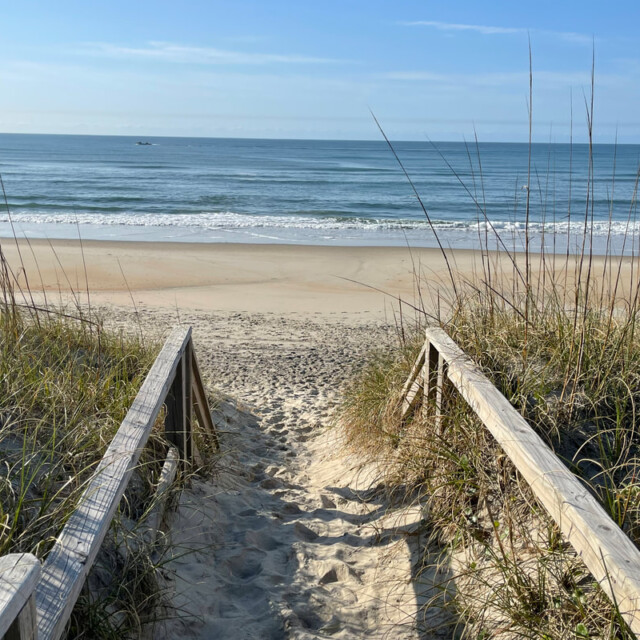 Wooden boardwalk with railings leads through grassy dunes to a sandy beach and calm ocean under a clear sky.