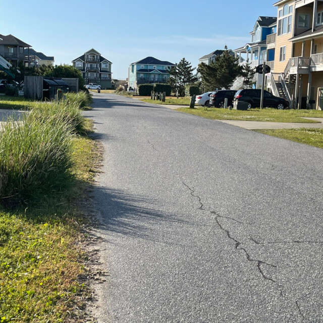 Paved residential street with beach houses, parked cars, and grass under a clear blue sky.