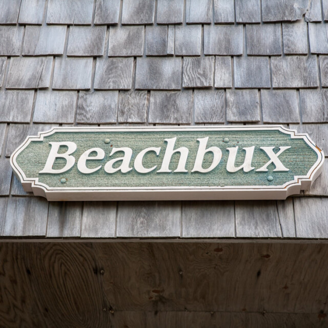 A green sign with the word “Beachbux” mounted on a wooden shingle wall.