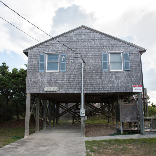 Wooden beach house on stilts with a “For Rent” sign and driveway underneath, surrounded by trees and grass.