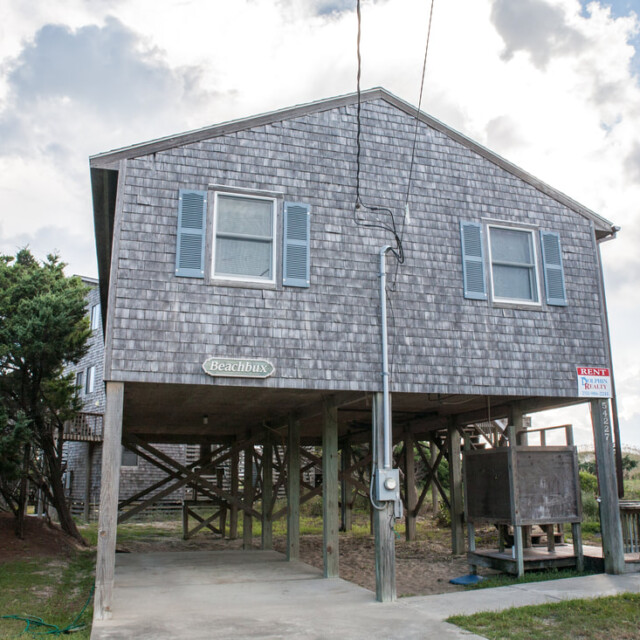 A gray, elevated beach house with blue shutters and a “For Rent” sign, sitting above an open parking area.