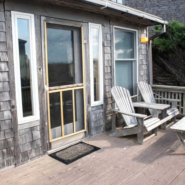 Wood deck with two Adirondack chairs, a bench, and a screen door leading into a weathered shingle-sided house.