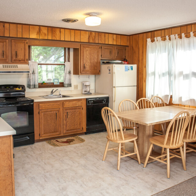 Kitchen with wood cabinets, black appliances, white fridge, wood dining table, six chairs, and a window with white curtains.