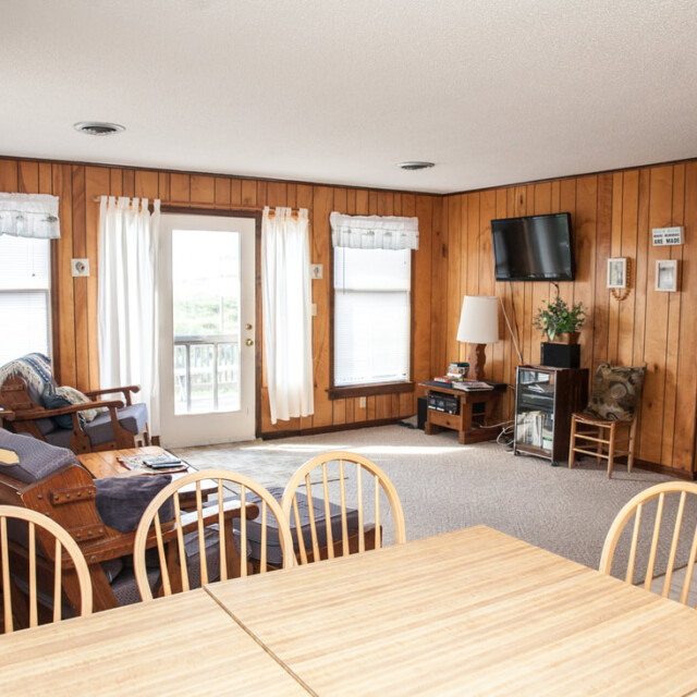 Bright living and dining area with wood-paneled walls, simple furniture, and windows letting in natural light.