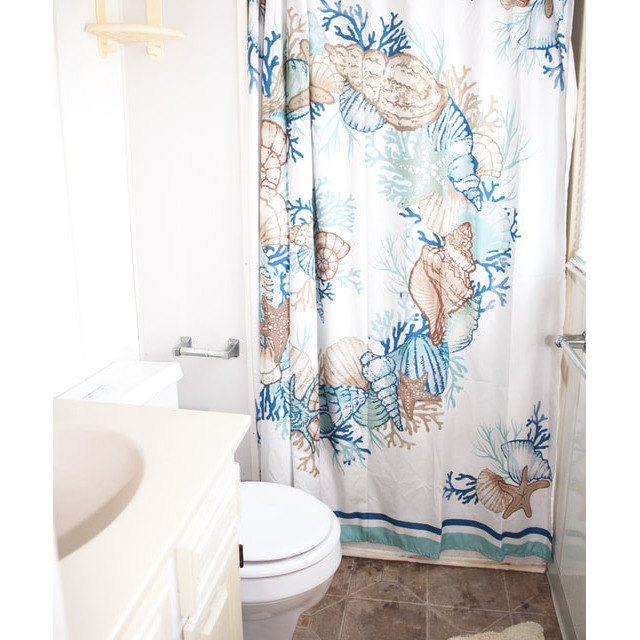 Small bathroom with seashell-patterned shower curtain, white toilet, beige vanity, and tan rug on tiled floor.