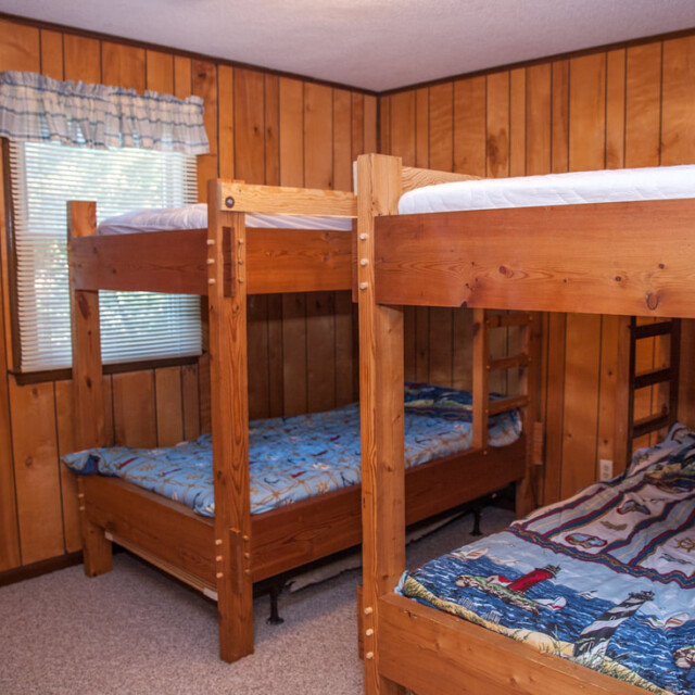 Wood-paneled room with two wooden bunk beds, nautical-themed bedding, carpeted floor, and a window with a valance.