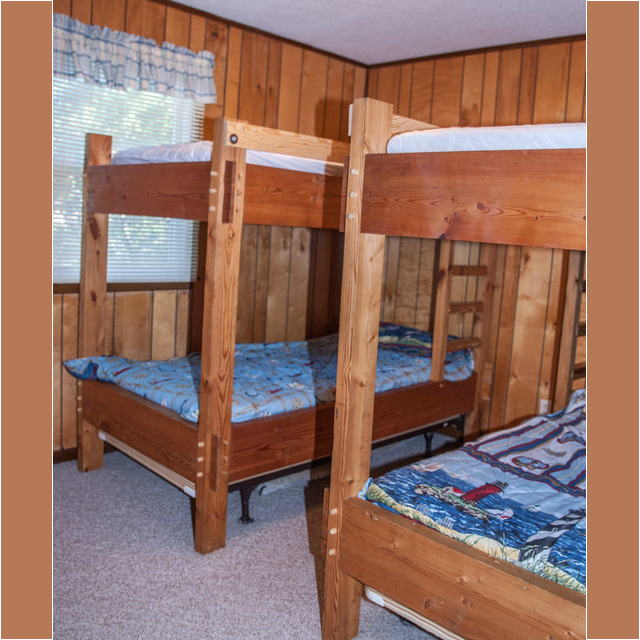 Two wooden bunk beds with blue bedding in a small room with wood-paneled walls and a window with a curtain.