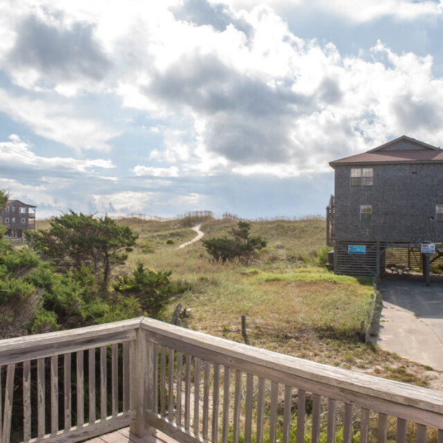 Wooden deck overlooks grassy dunes, a path leading to the beach, and two houses under a partly cloudy sky.
