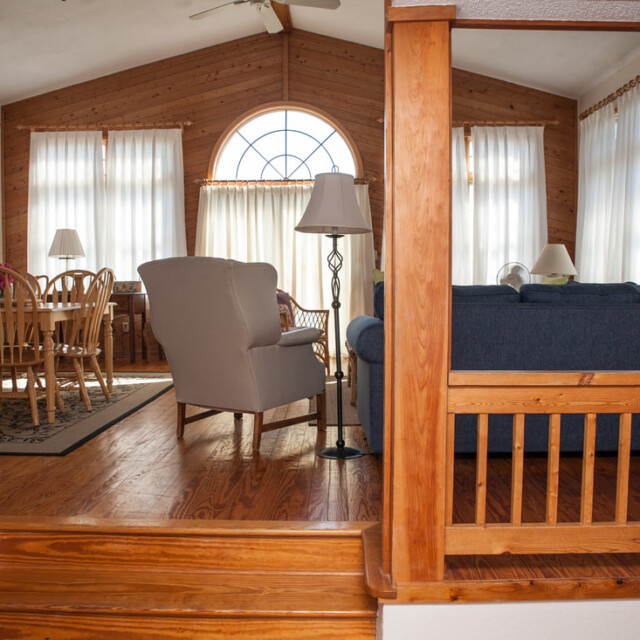 Sunlit living and dining area with wood floors, blue sofa, beige armchair, and wooden dining table near large arched window.