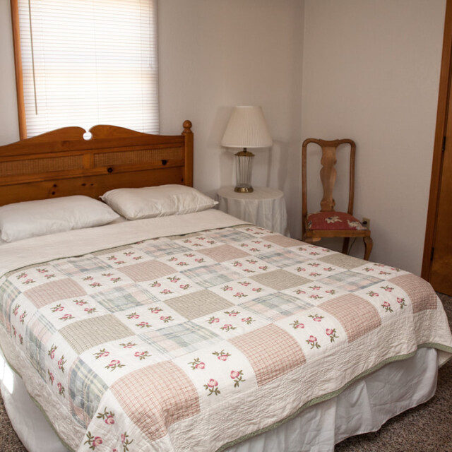 A neatly made bed with a patchwork quilt, two pillows, and two bedside tables with lamps in a simple bedroom.