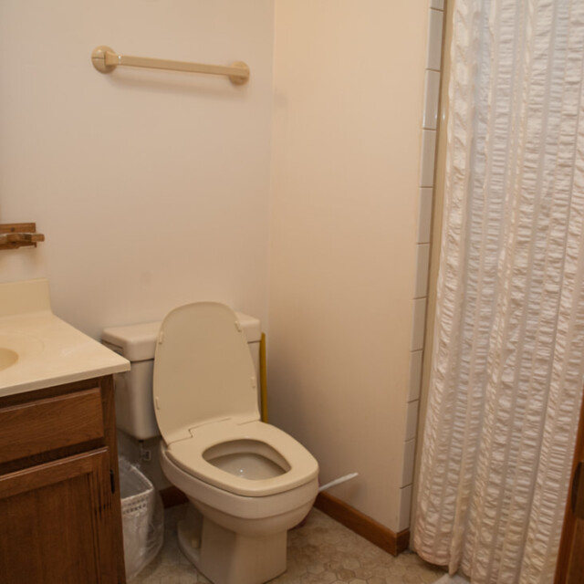 Small bathroom with a toilet, wooden vanity, wicker mirror, towel bar, and white shower curtain.