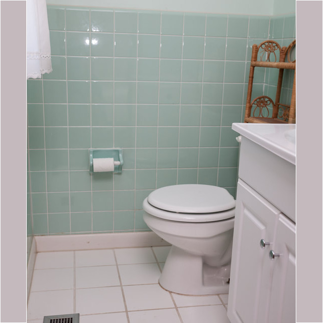Bathroom with mint green tiled wall, white toilet, toilet paper holder, vent, and white vanity cabinet.