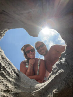 Two people in swimsuits smile through a heart-shaped hole in sand with the sun shining in the background.