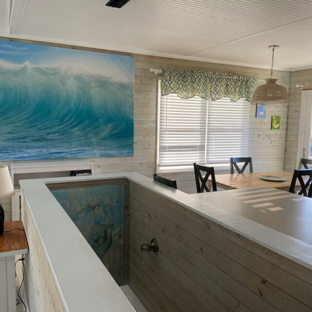 Sunny dining area with wooden walls, a large ocean wave painting, and a staircase with a glass railing.