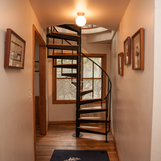 A narrow hallway with wooden floors leads to a black spiral staircase and a window with a view of trees.
