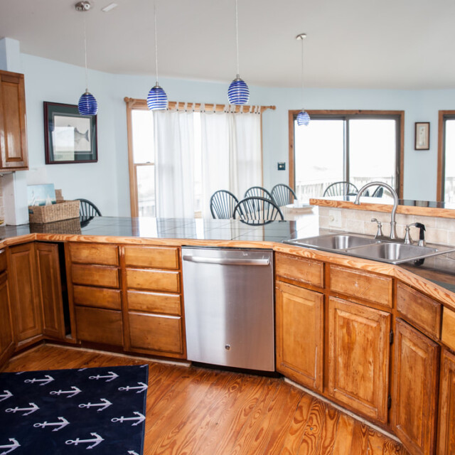 Spacious kitchen with wooden cabinets, stainless steel dishwasher, double sink, and bar seating area facing large windows.