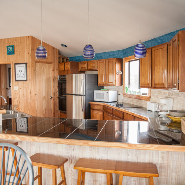 Kitchen with wooden cabinets, stainless steel appliances, black tiled counter, and three blue pendant lights over a bar.