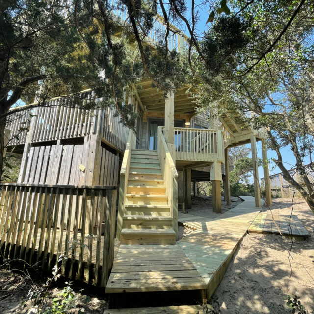 Two-story wooden treehouse with stairs and railings, surrounded by trees and sand under a clear blue sky.