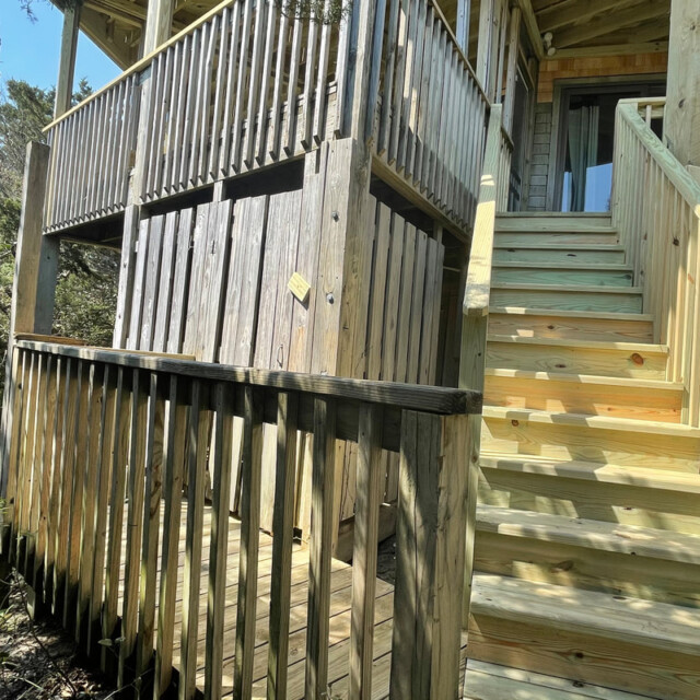 Wooden stairs and railings leading to a raised deck; lower enclosed area beneath the deck; surrounded by trees.